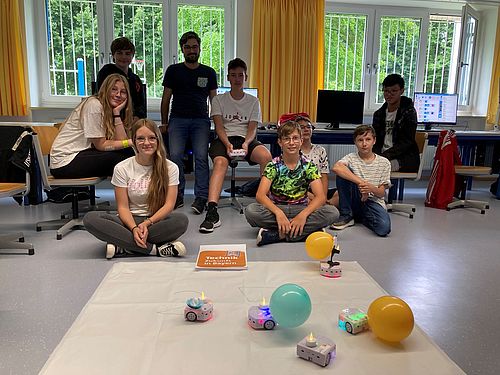 Happy Birthday zu 25 Jahren Technik – Zukunft in Bayern im Vordergrund schreiben 2 Thymio-Radroboter eine 25; Schülerinnen sitzen im Hintergrund