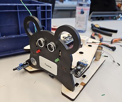 Man sieht ein Modell des Roboters. Es sieht von vorne aus wie eine Maus und hinten sind einfache Holzbretter angebracht. 