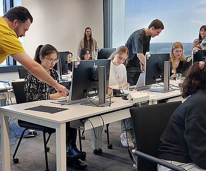 Es ist in einem Computerraum. Die Mädchen sitzen an den Tischen mit den Computern. Zwei Männer erklären zwei Mädchen etwas und deuten dabei auf den Bildschirm. 