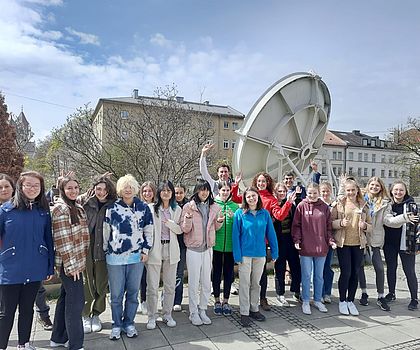 Die gesamte Gruppe Mädchen steht vor einem Satelliten und sie haben die Arme vor Freude in der Höhe. 